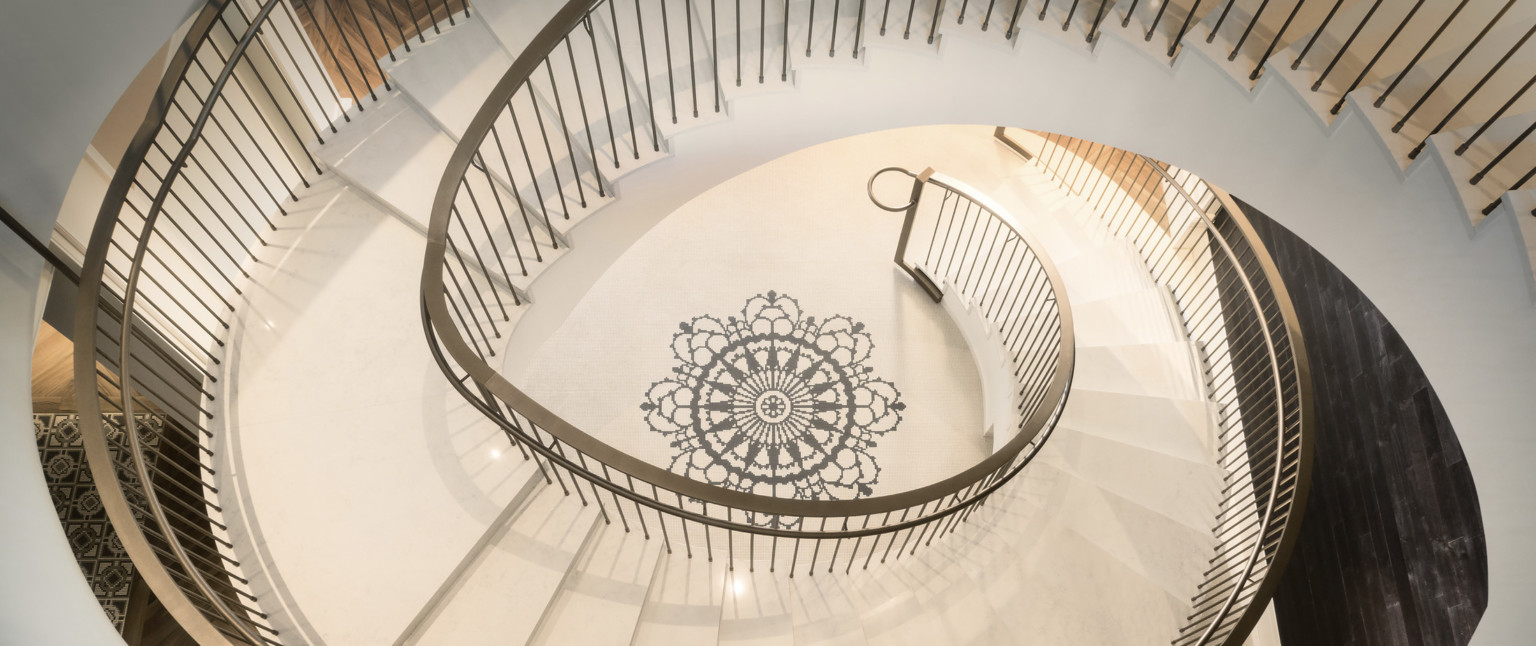 View from above of white spiral staircase with metallic railing. At the center of the floor in the spiral is a black mandala