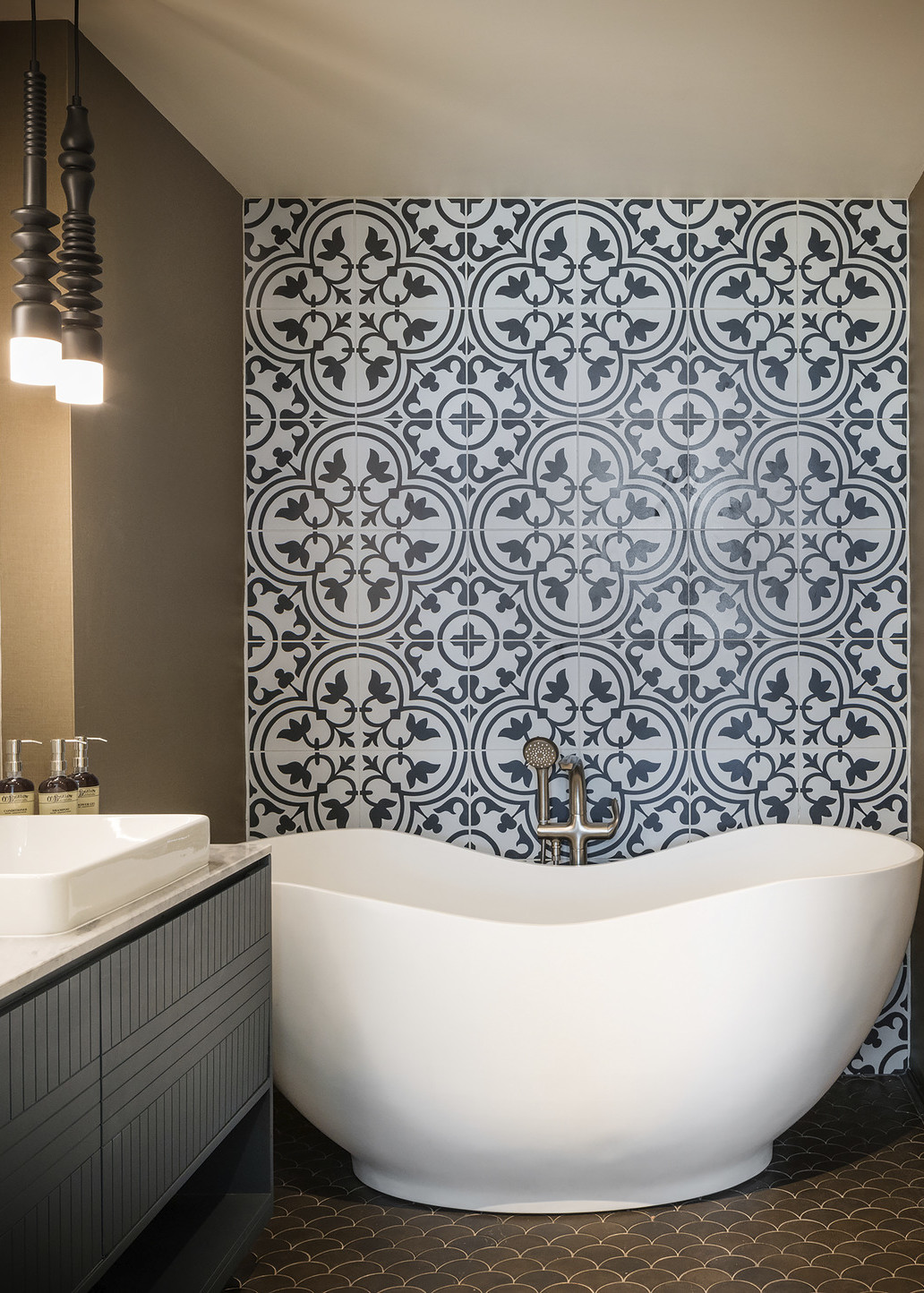 White curved tub in front of blue toned grey patterned tile wall in brown bathroom with sink and pendant lights to the left