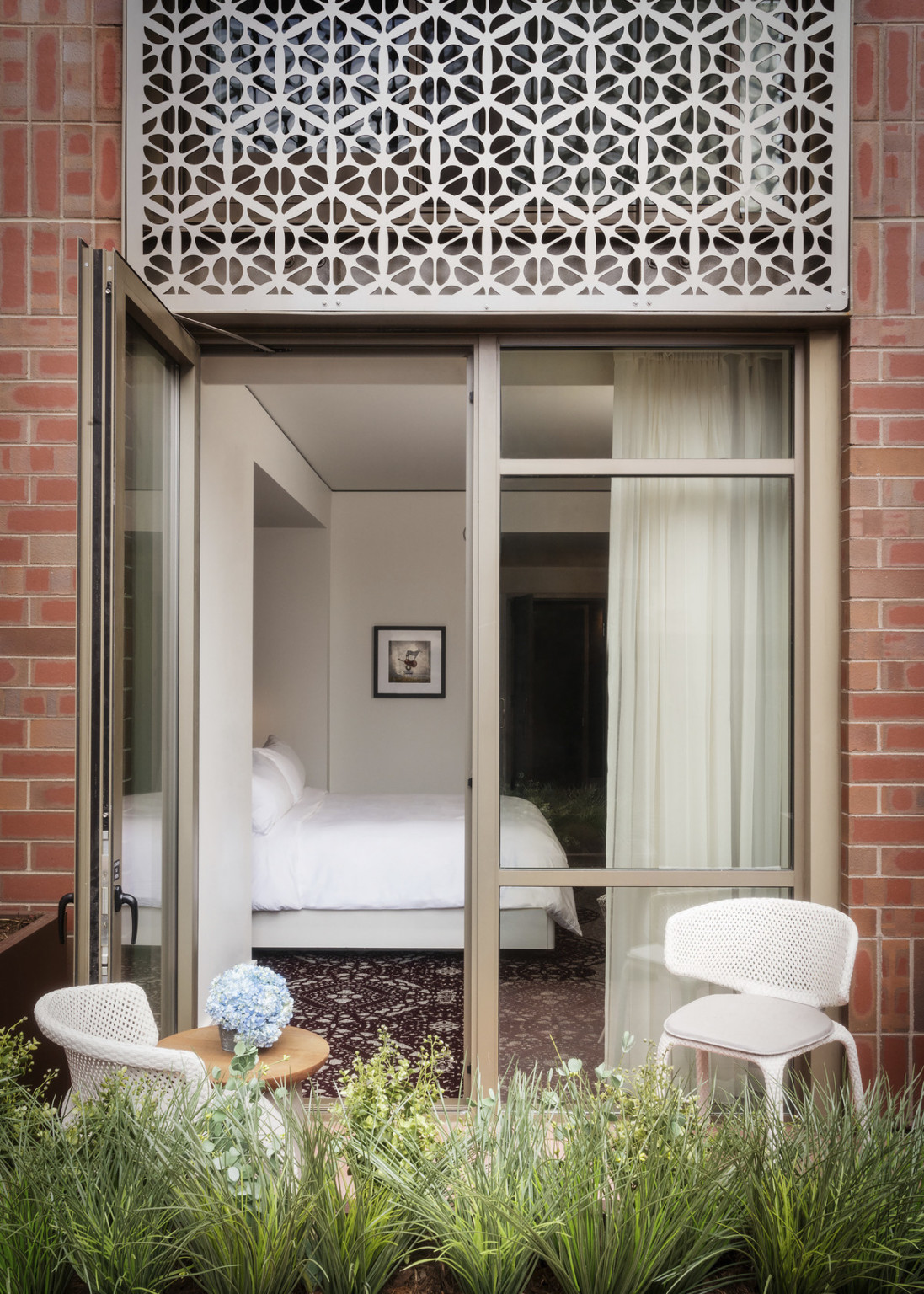 Balcony off guest room with planter in front of white chairs and small round wood table. Metal with cutouts over door