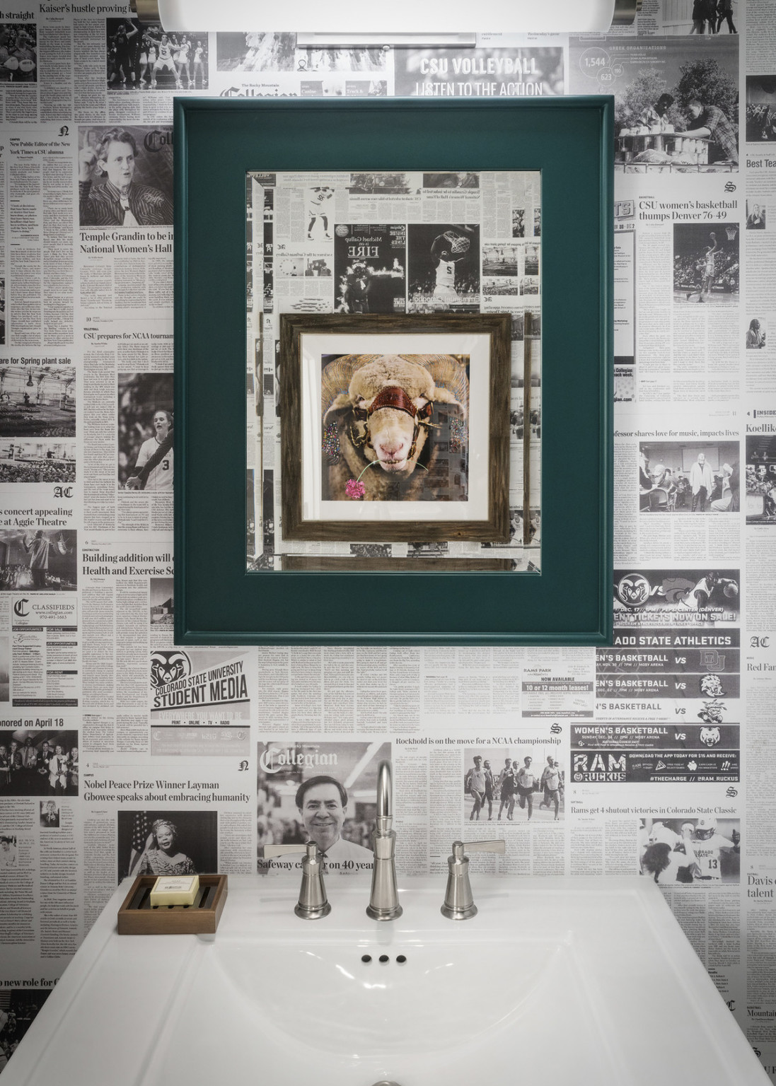 Framed picture within a green frame on wall with black and white newspaper wallpaper over white sink with silver faucets