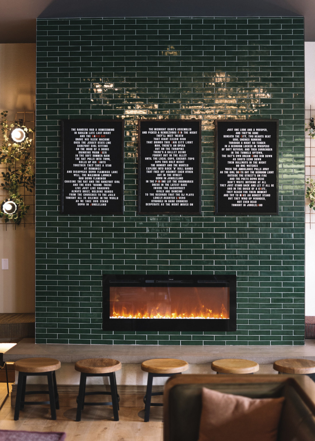 Green fireplace closeup with 3 panels of white words on black canvas. Stools in front of fire. White walls on side with art