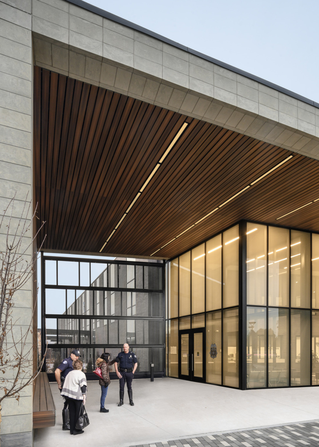 Building entry with double height windows, wood panel detail and lighting sit under stone overhang and down the wall to bench