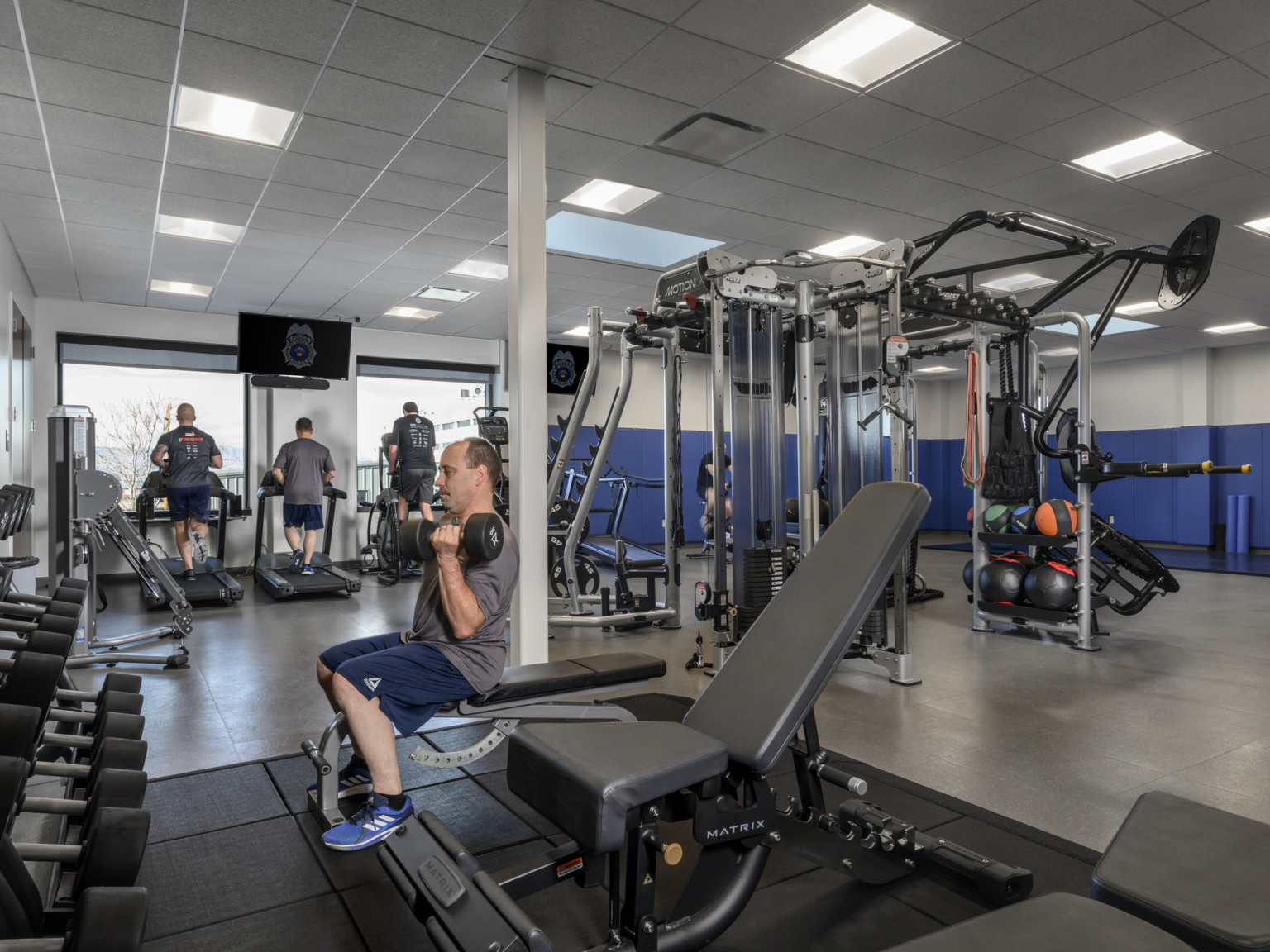 Gym weight room with police officers exercising under square white ceiling tiles and blue colorblocking on rear wall