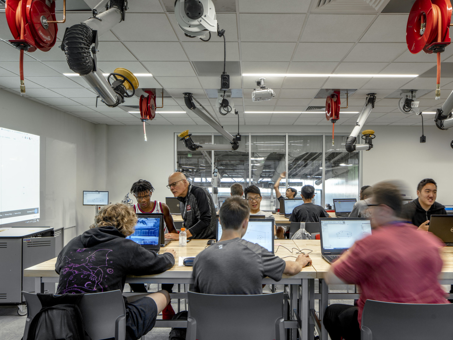 Classroom with students on computers at rows of wood tables. Equipment and power cords hang from the ceiling of white room