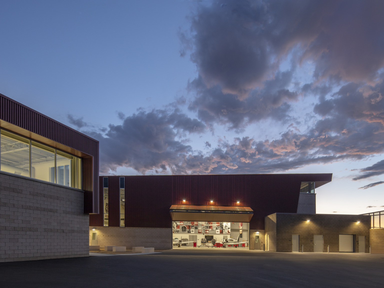 Stone building with textured red wrap facade illuminated from within in evening. Garage door opened at center to airplanes