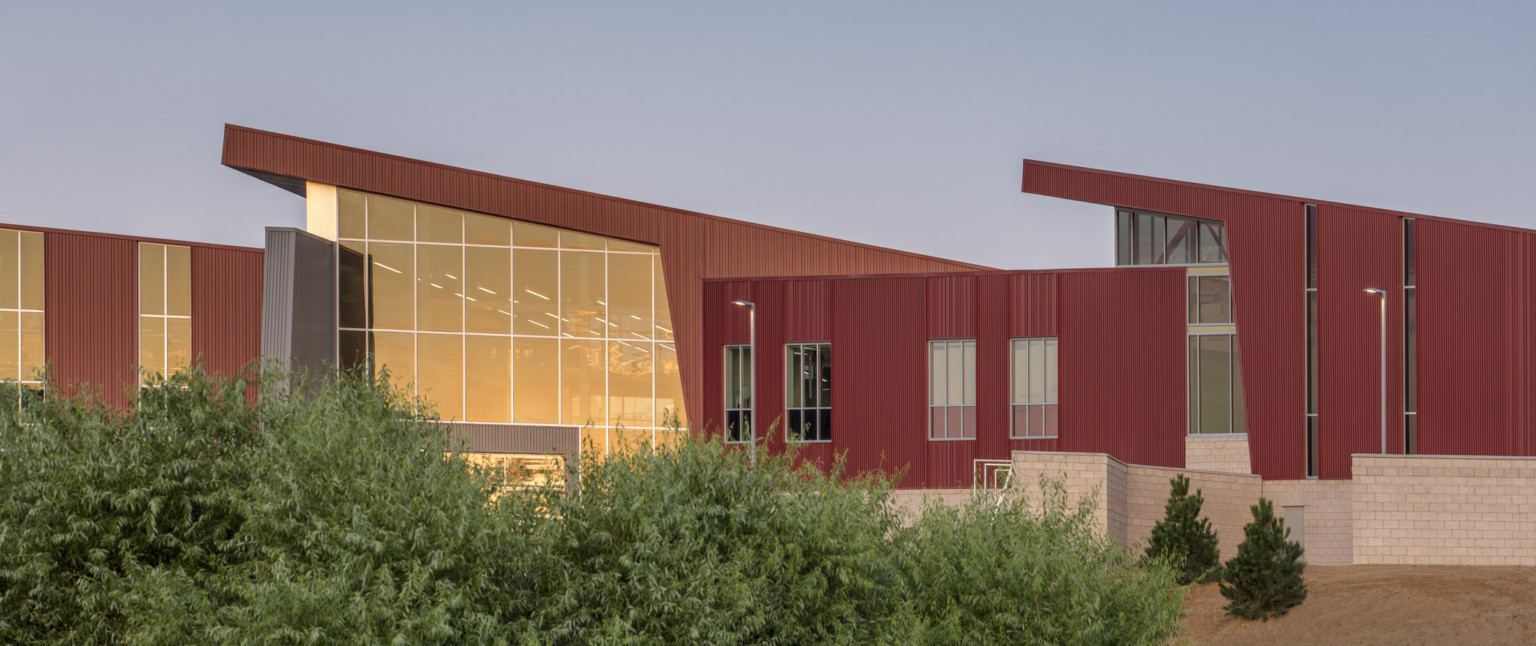 Stone base on building below textured red panel wrapped facade with angled roofs. Angled double height glass entrance