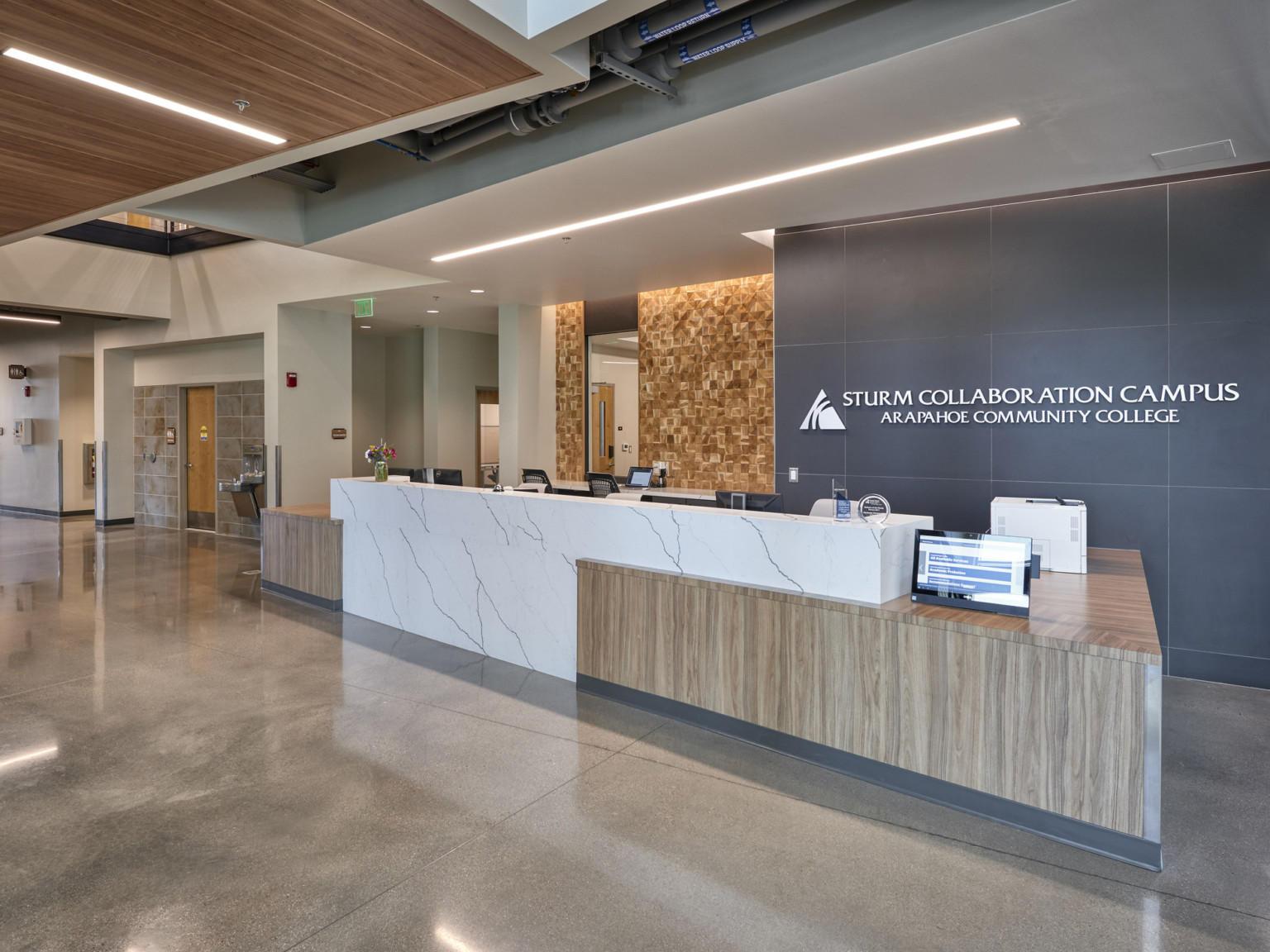 Reception area with white marble desk with wood accents to side in front of stone wall with Sturm Collaboration Campus sign.