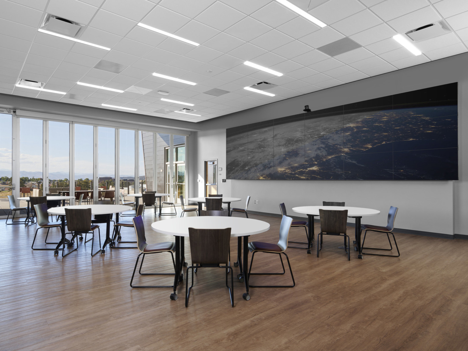 Round tables with black chairs in grey room with floor to ceiling windows. Long screen on right wall, white panel ceiling