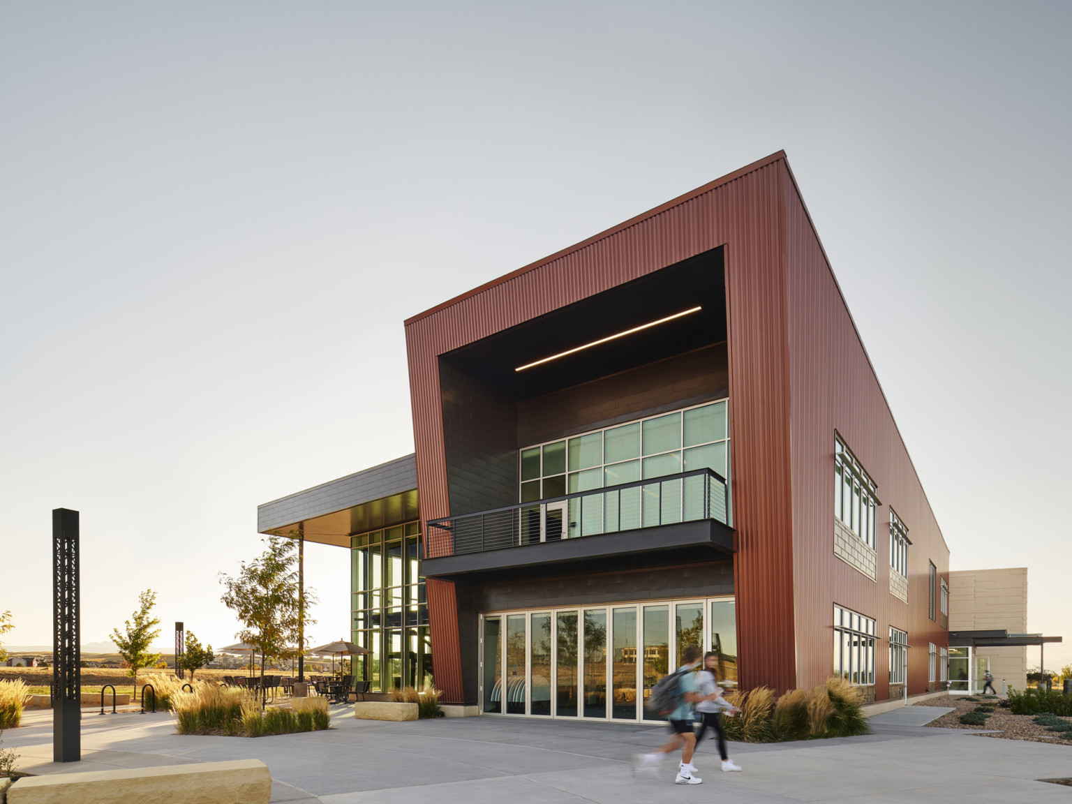 Building with angular dark awning extending above double height glass façade. Tables and umbrellas outside, with trees