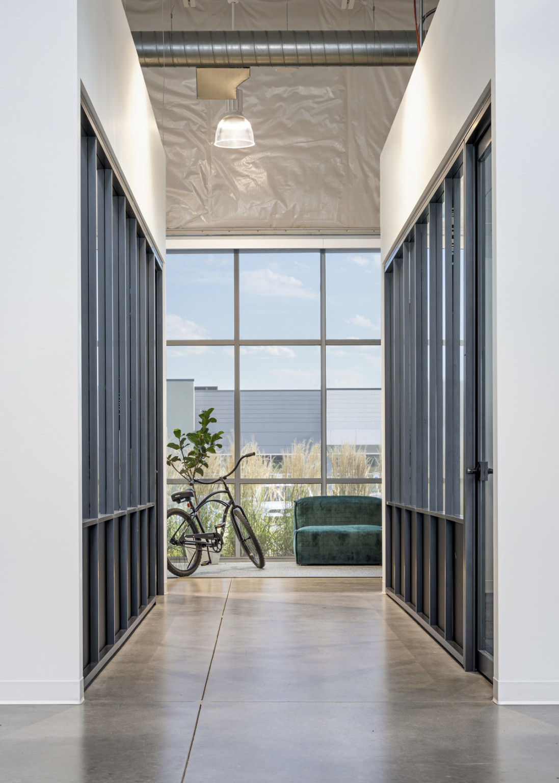 White hallway with windowed rooms leading to tall window lined from outside with plants along bottom. Exposed duct overhead.