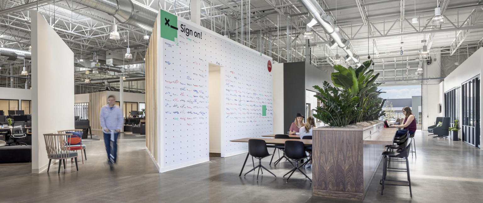 Tables in front of wood half wall with plants on top and counter attached. White board wall to left. Open garage door window