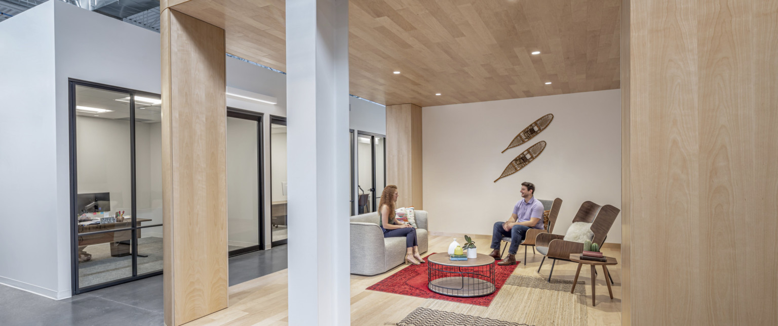 Wood alcove opposite glass walled offices in white hallway. Decorative snowshoes on wall above seating area framed by columns