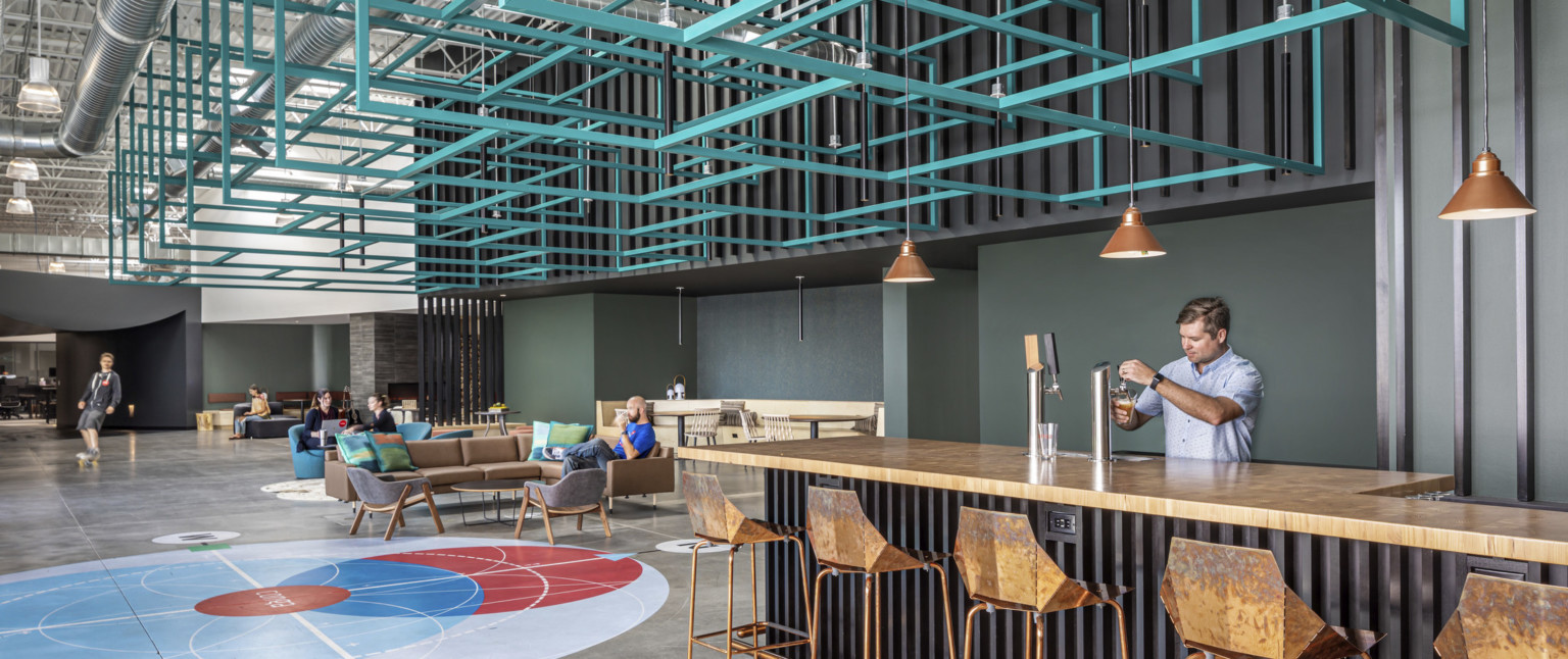 Counter with beer taps in large double height hallway lined with seating. Above, blue metal angular sculpture hangs