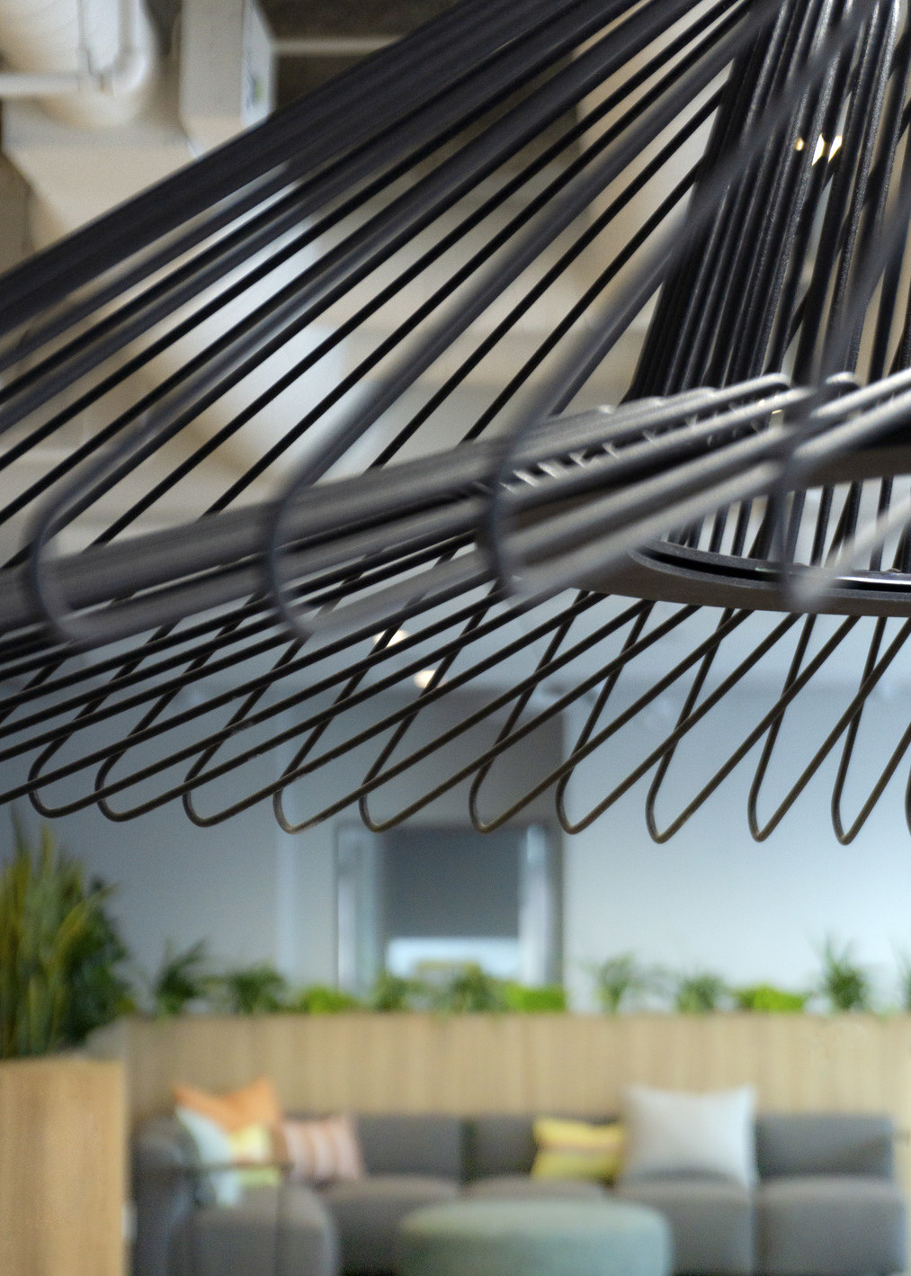 Closeup of black wire pendant lamp shade. In background, couch with wood half wall with plants behind