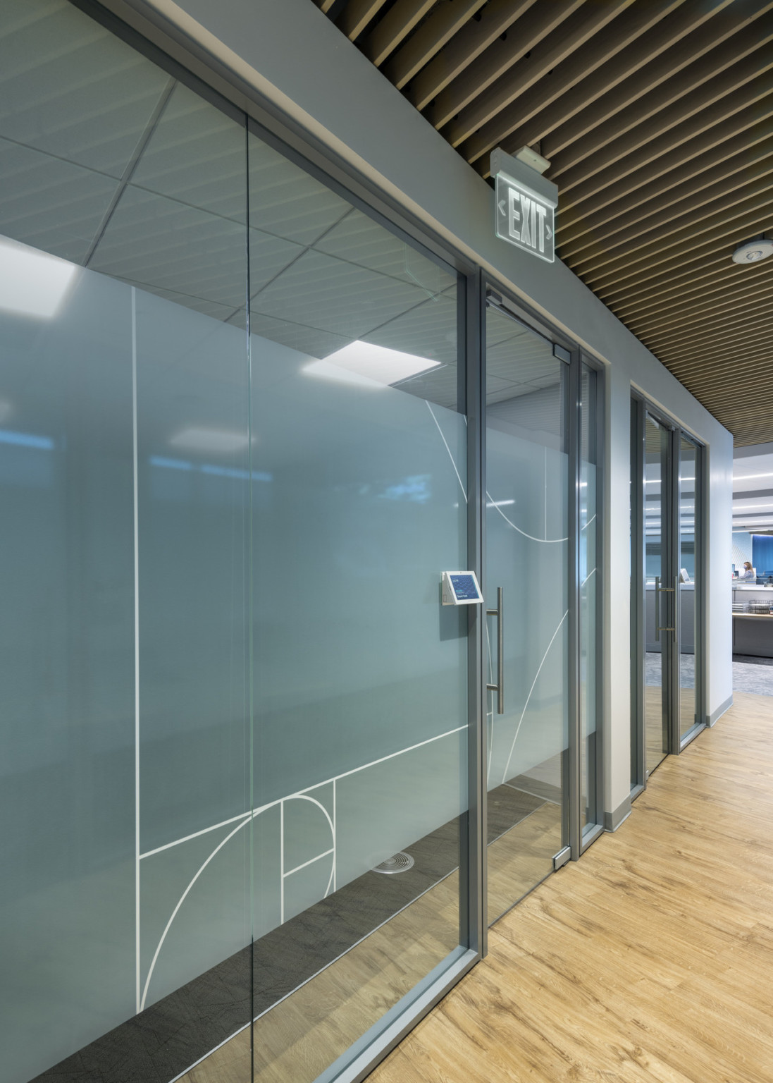 Wood slat ceiling in hallway with glass walled rooms to left with translucent bar and abstract design across center