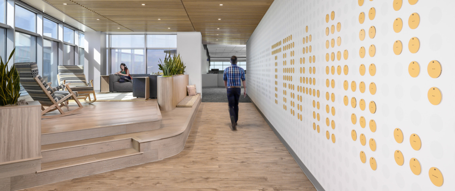 Wood slat ceiling over hallway, mezzanine with floor to ceiling windows and seating to left. Wall, right, with circle details