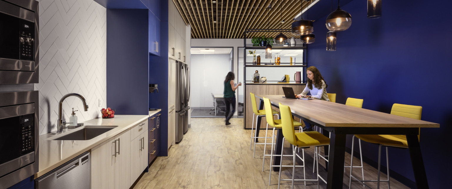 Kitchenette with dark blue walls and light wood cabinets. Recessed white tile over counter, left. High top table to the right