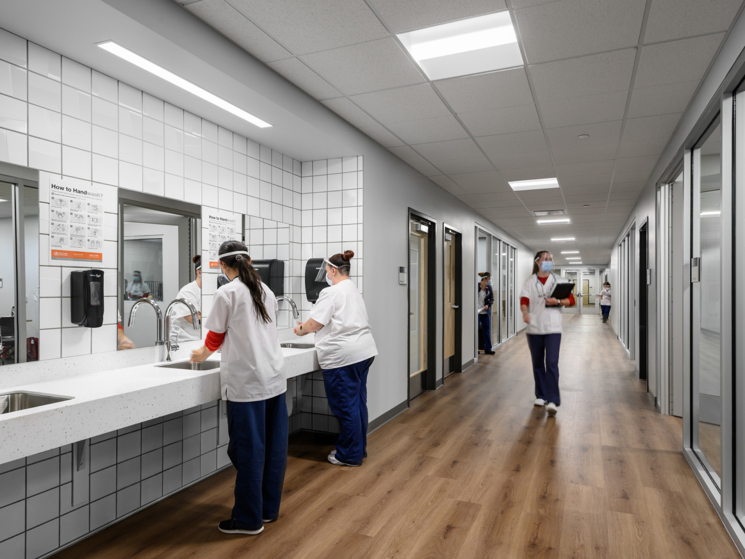 White hallway with hardwood floors. Glass walled rooms to right, section of counter space left with sinks and tile backsplash