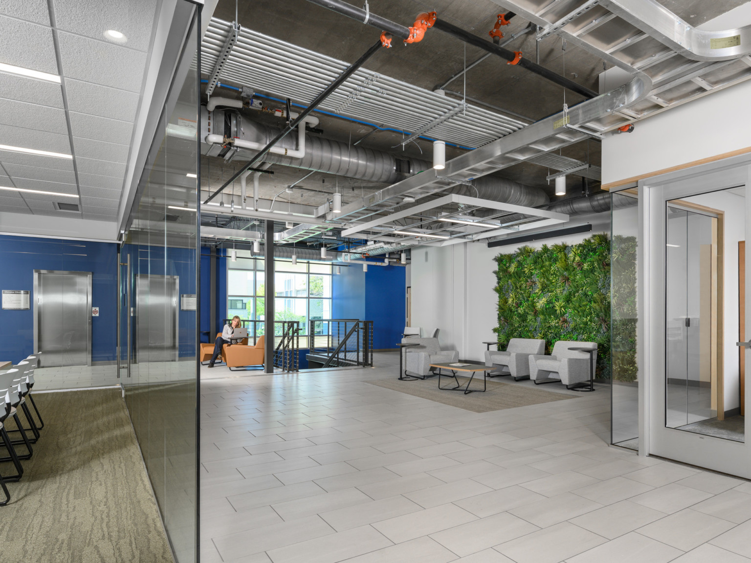 Hallway with blue accent wall by elevators, seating area in front of living wall detail right. Transparent work space left