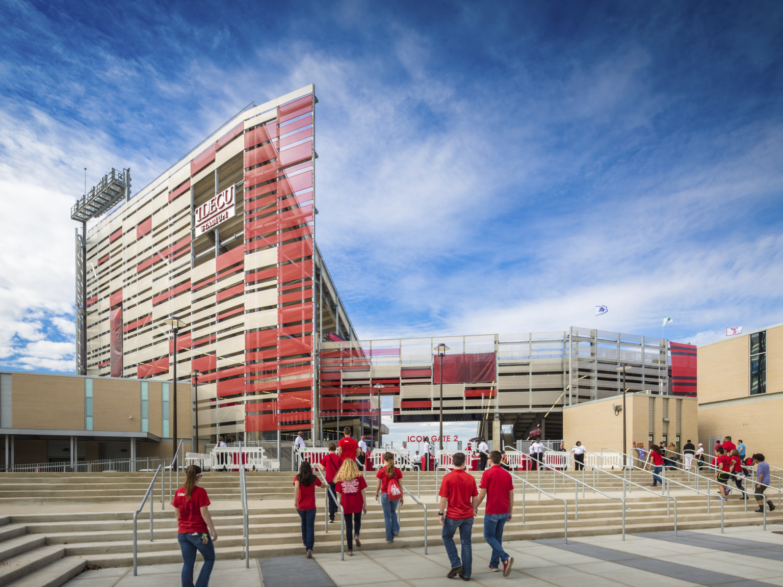 Stadium with red and white sunscreen wrapped angularly along facade. Stairs lead to entrance, center, between stone buildings