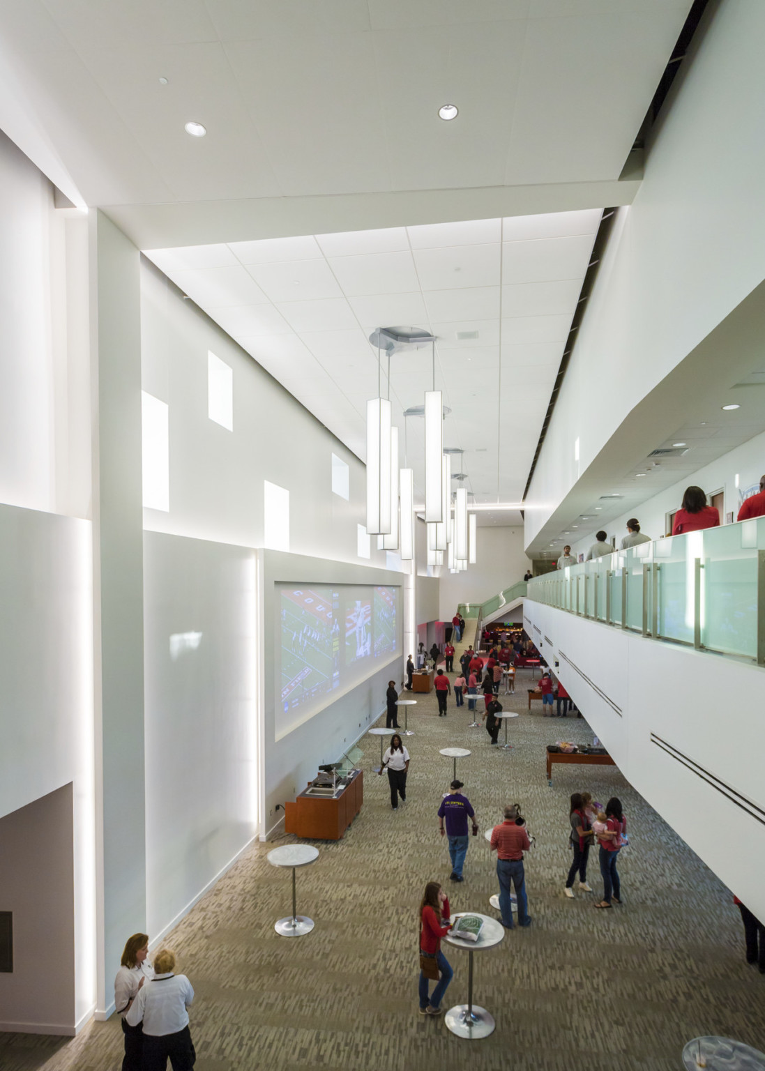 Interior double height room with tables on 1st floor, 2nd floor walkway with glass rail, right. Sculptural chandeliers hang