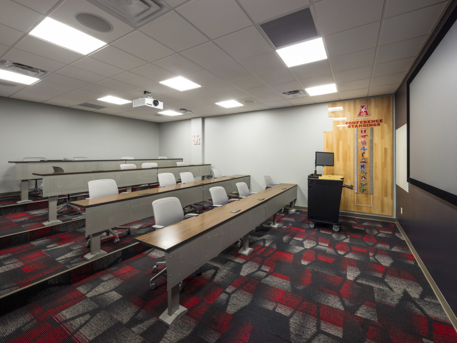 Long tables with rolling chairs on stepped incline face podium and screen in white room with red patterned carpet