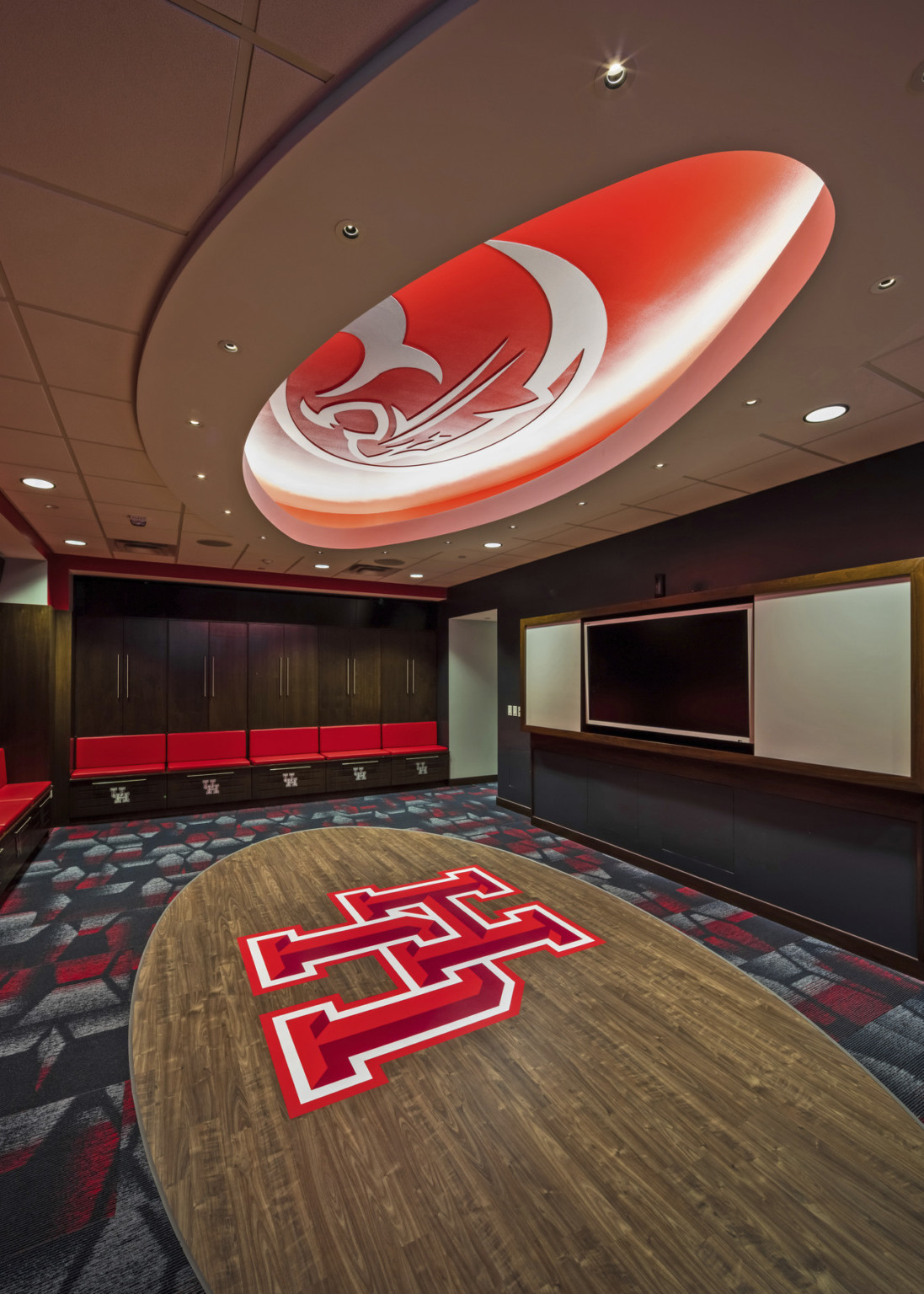 Locker room with wood lockers and red seats. Oval floor panel with UH logo mirrored by recessed ceiling with mascot image