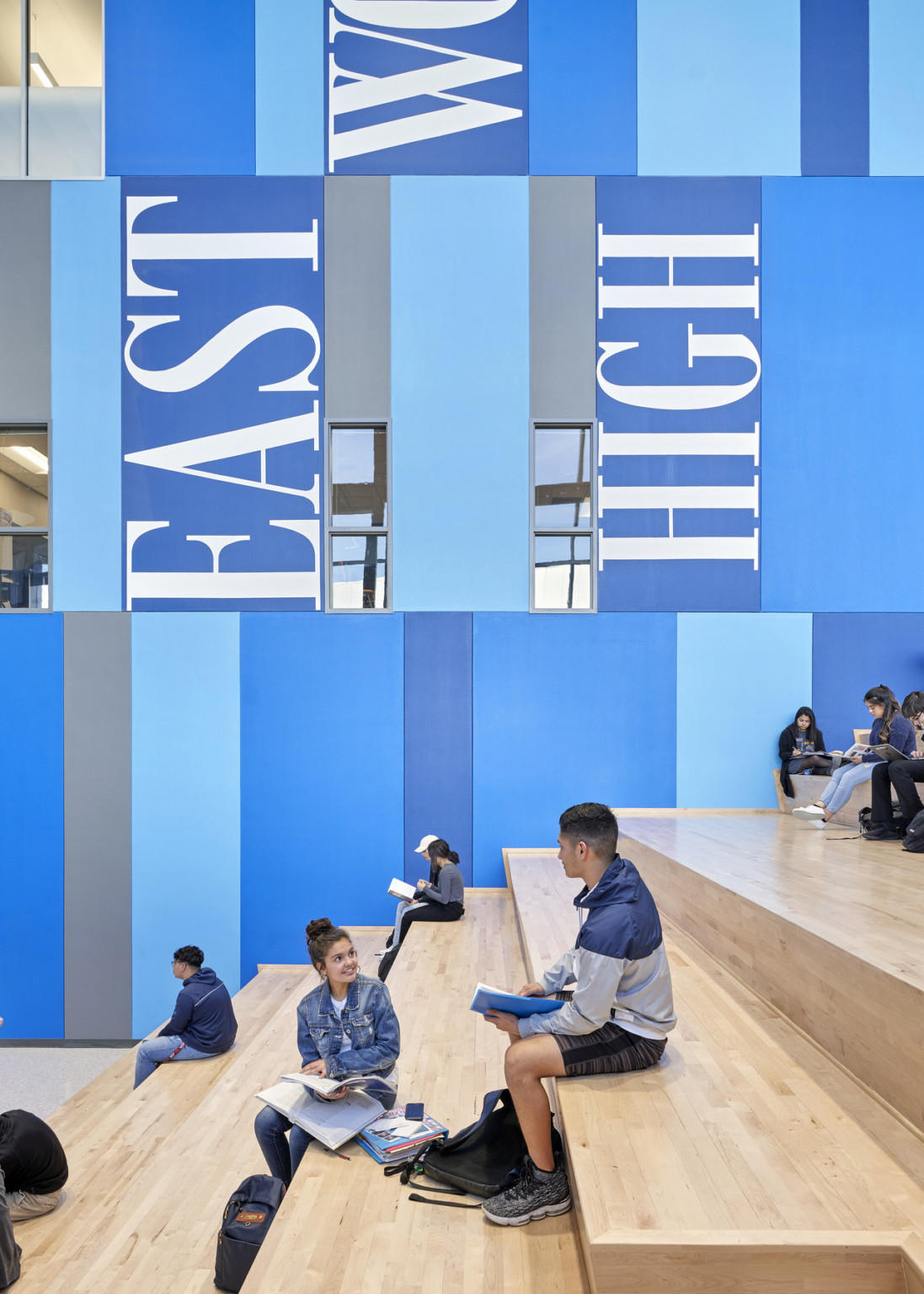 Wood step bench seating along a blue colorblock wall with Eastwood High written on it. Thin windows to room on other side