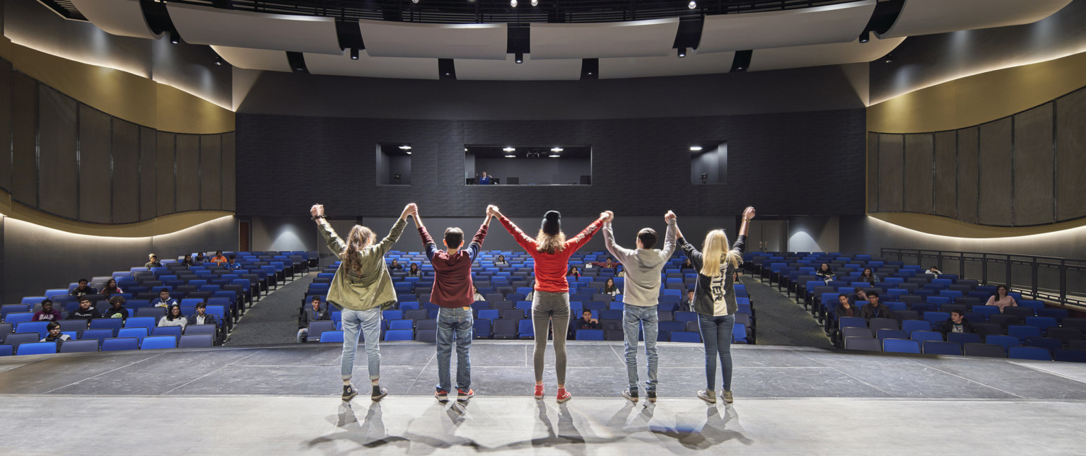 Students stand on stage facing audience. Curved acoustic panels above. Wood details arch along side walls with lights behind