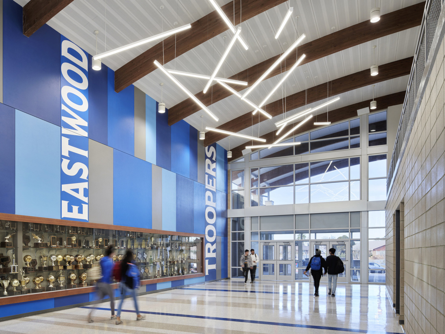 Hall with curved white roof and exposed wood beams. Bar lights hang abstractly. Right is blue mural with recessed trophy case