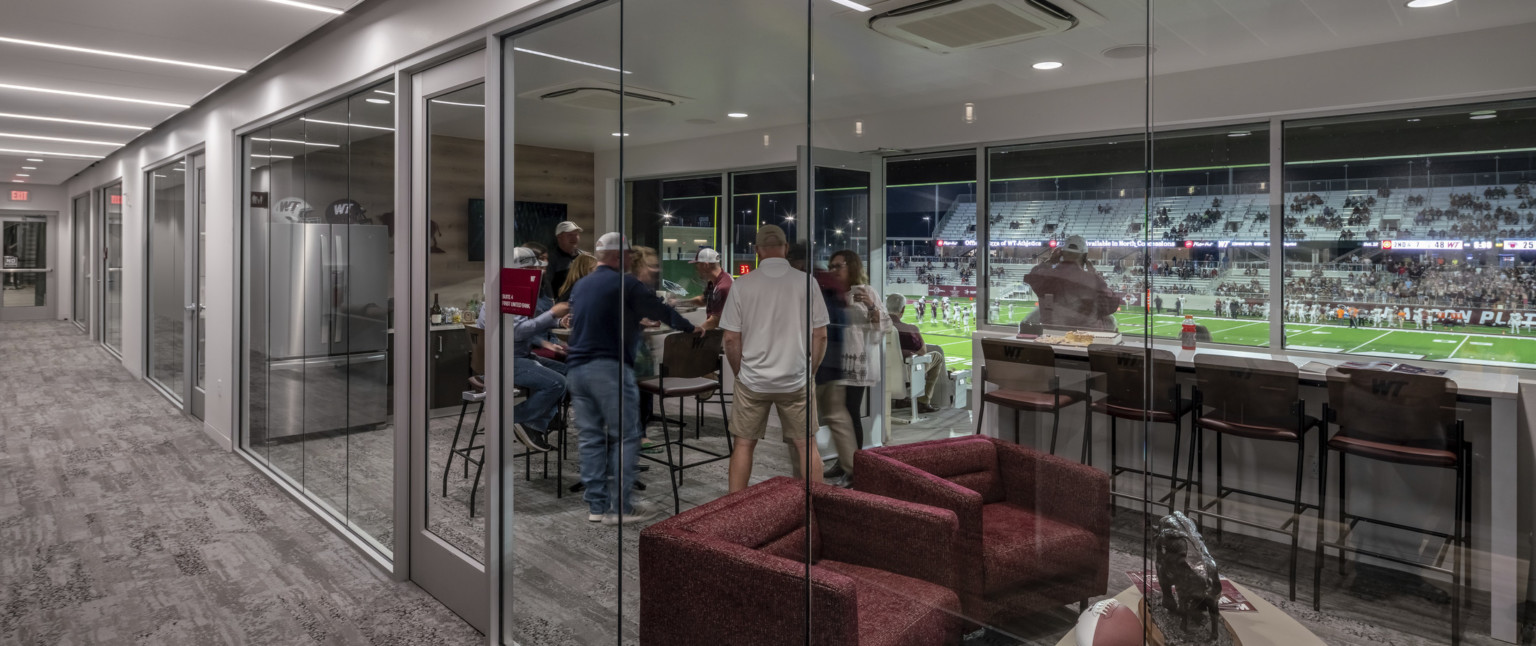 Private suite with glass walls to hallway and field. Inside, armchairs and tables, refrigerator and counter space on far wall