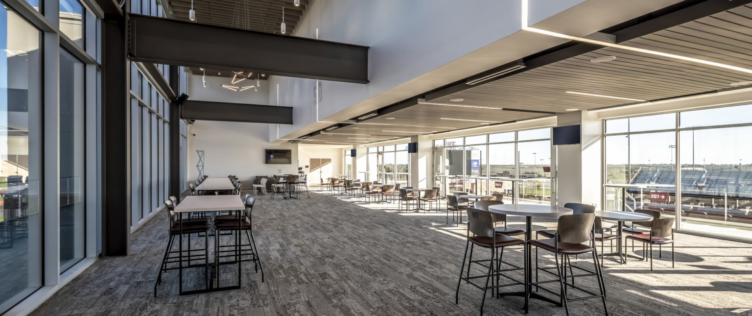 Double height room with black exposed beams and drop ceiling to the right above floor to ceiling windows. Tables in the room