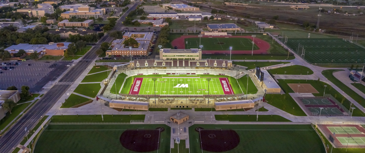 Aerial view of stadium with WT logo in white at field center. Soccer field behind and basketball courts right, buildings left