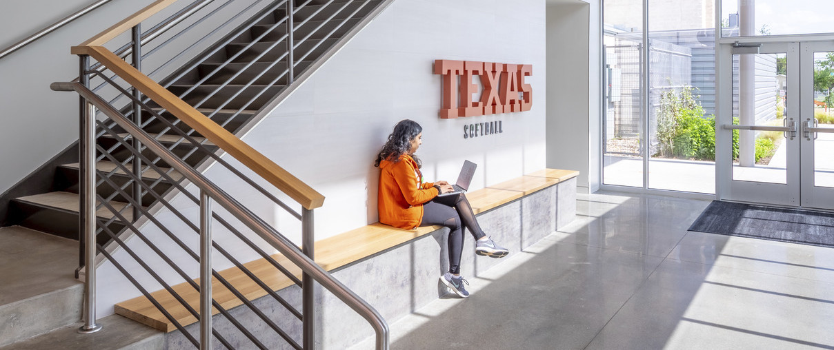 Wood bench along white wall of stair with Texas Softball sign. Glass wall and door leading to exterior walkway