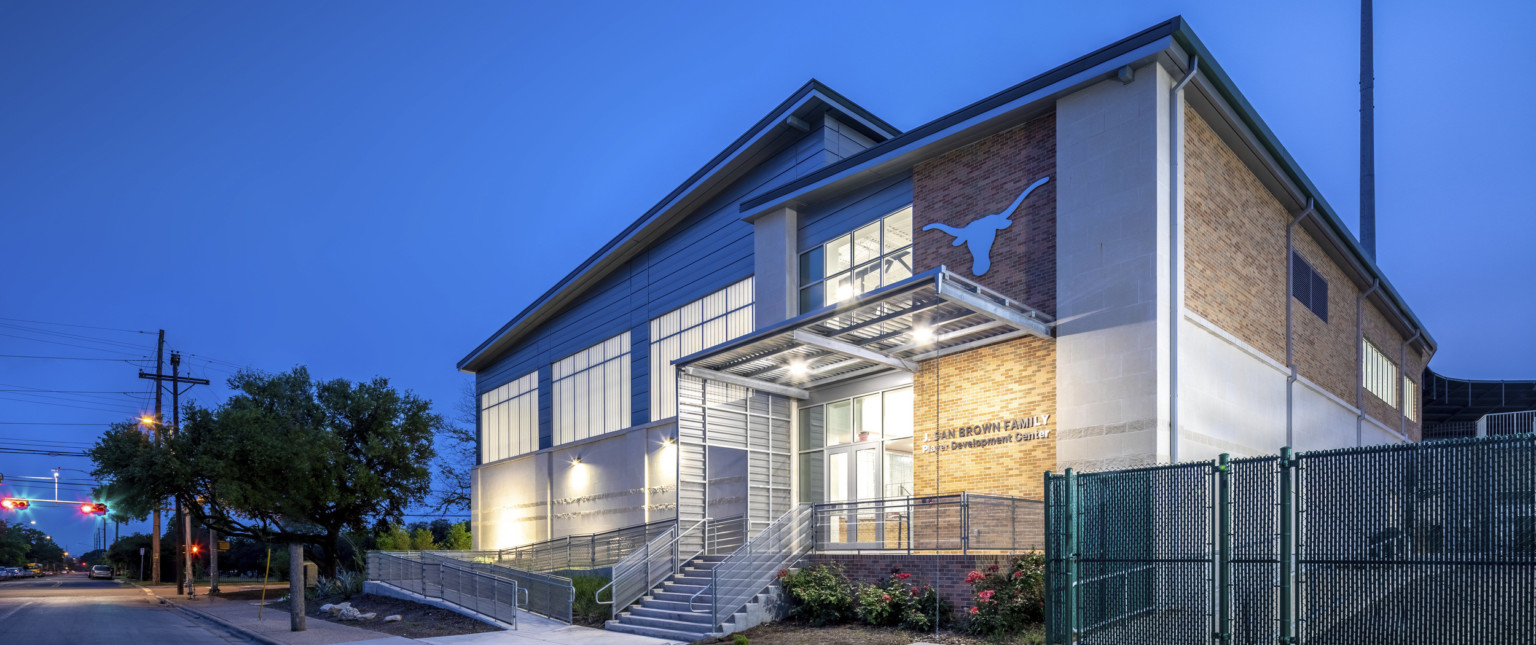 Corner view of brick building with white wrap facade. Connected white panel building with angled roof, left. Sheltered entry