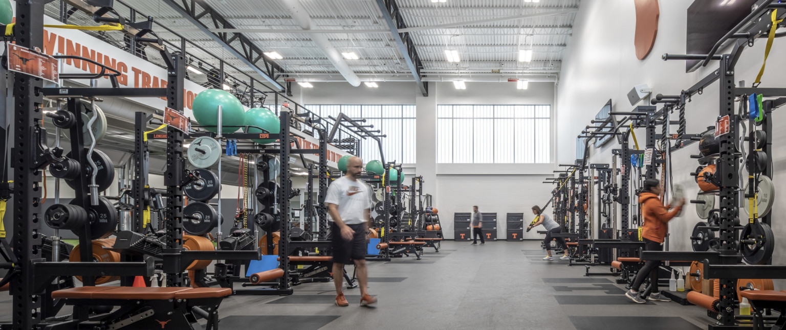 Training room with weight equipment along both walls. Double height white room with more equipment on 2nd floor balcony