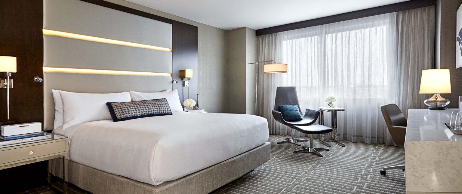 Guest room with white recessed lighting in headboard. A large window, grey chair and foot rest, and round lamp in grey room