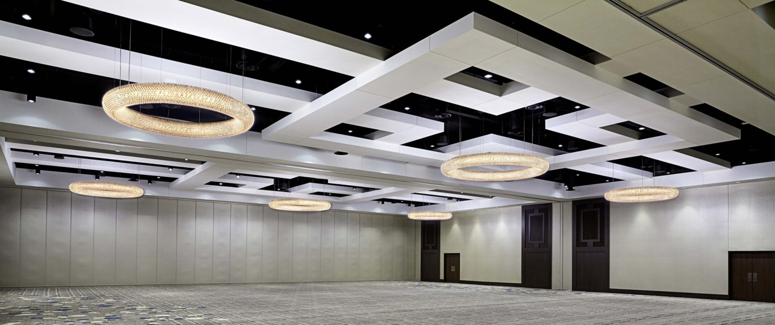 Hotel ballroom with large interlocking white geometric drop ceiling details and round illuminated chandeliers in cream room