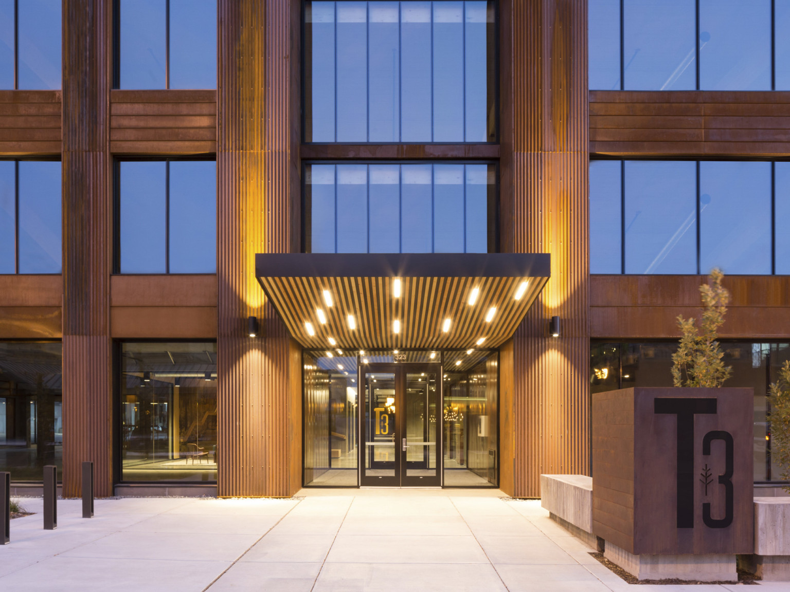 Front entrance to a mass timber building with glass door. Awning with recessed lighting in wood slat plans. T3 sign, right, mass timber buildings, t3 design, mass timber high rise, t3 construction, t3 partners