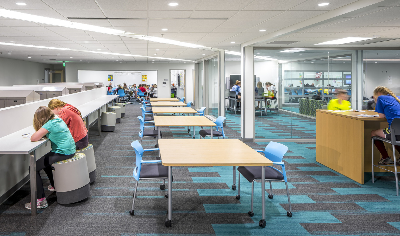 Long white room with white counter and stools, left. Tables with blue chairs, right. Glass walled learning spaces far right