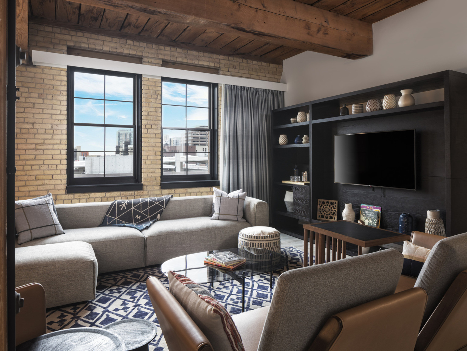 Sitting area with black wood entertainment center with shelves and television. Exposed brick wall under wood ceiling, adaptive reuse hotel