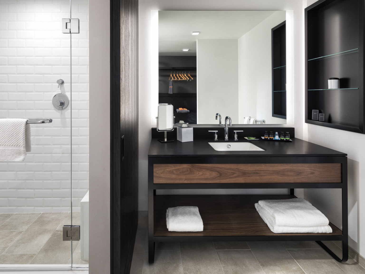 Bathroom with white tiled walk in shower left of black and wood sink. Black accent wall left of sink and black shelves right