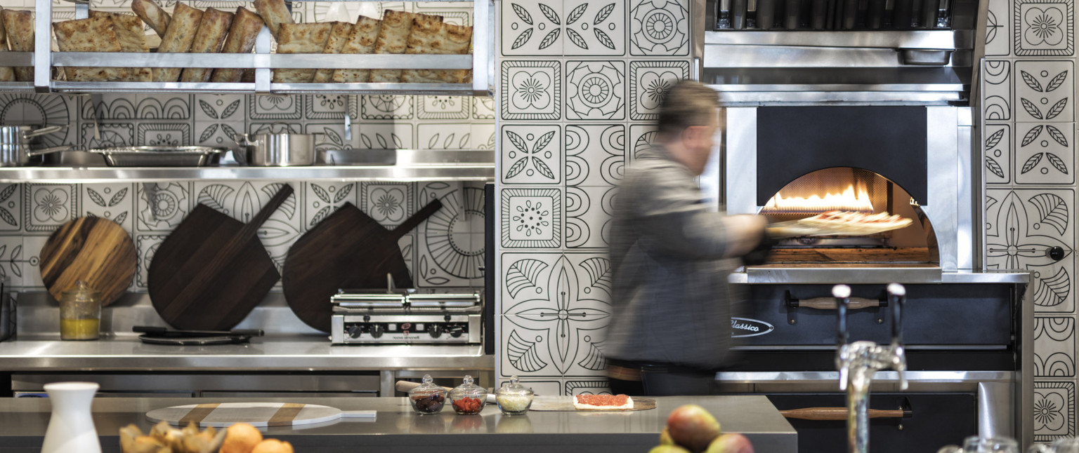 Pizza being placed into oven in kitchen with black and white floral tiles. Stainless steel counters and shelving with bread, adaptive reuse hotel