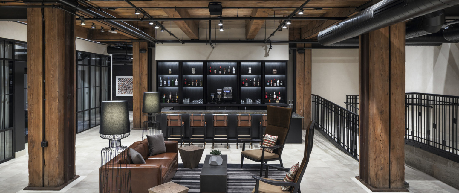 Comfortable seating in front of black bar in white room with wood ceiling, beams, and columns. Black metal railing to right, adaptive reuse hotel
