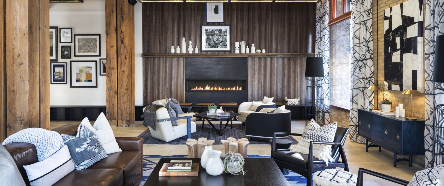 Seating area with arm chairs and couches in front of wood fireplace in white room with large curtained window, adaptive reuse hotel