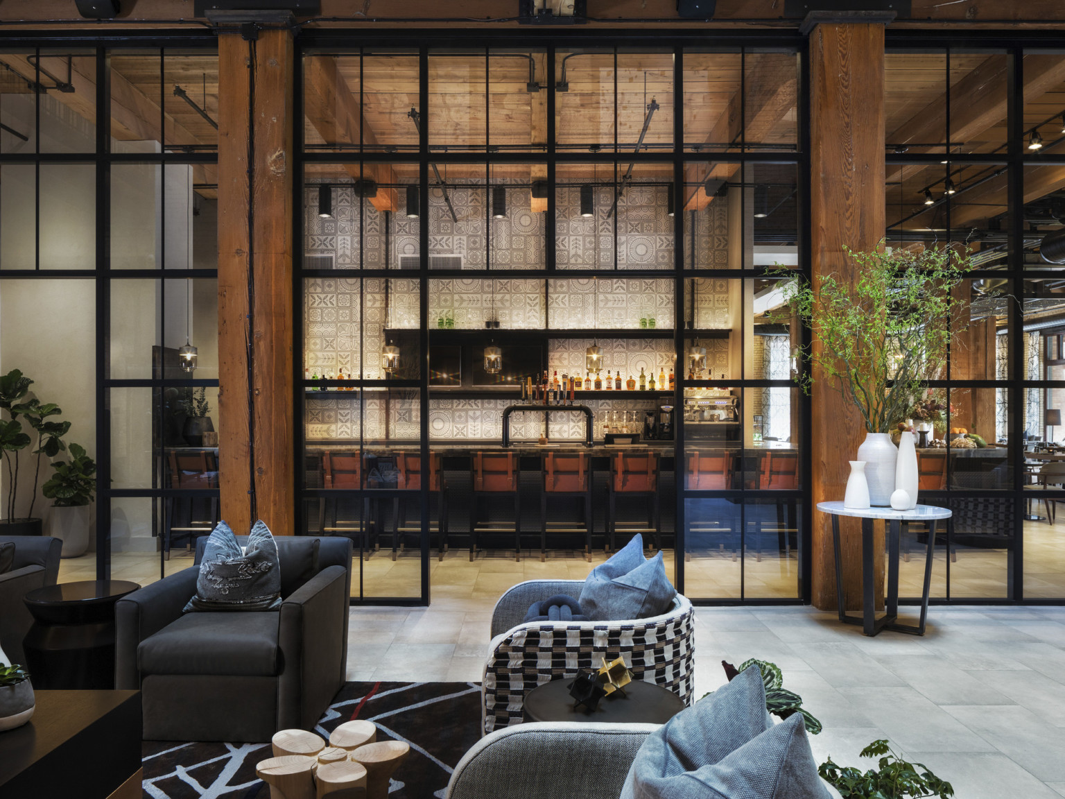 Seating area in room with double height windows looking to white geometric walled bar in next room. Wood columns and ceiling