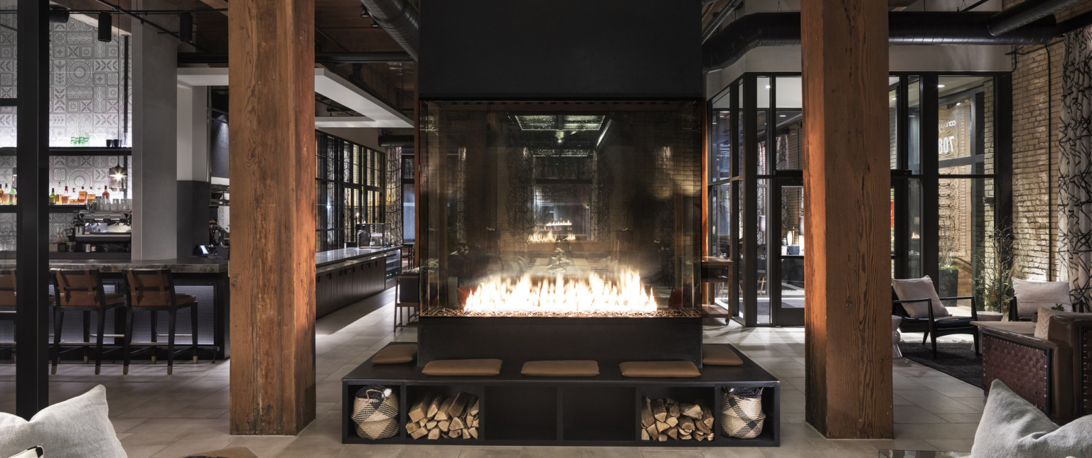Fireplace elevated on black bench with under seat storage between wood pillars in bar with floor to ceiling windows, adaptive reuse hotel
