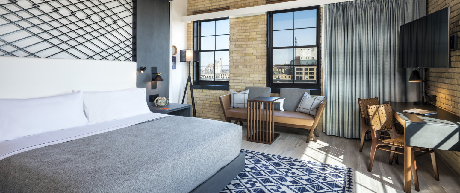 Guest room with exposed brick wall. Bed with white headboard against black metallic grid anchored to grey wall accent, adaptive reuse hotel