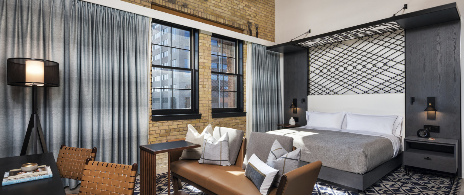 Bedroom with geometric black metal design behind wall and folding over top of bed to form canopy. Left, exposed brick wall, adaptive reuse hotel