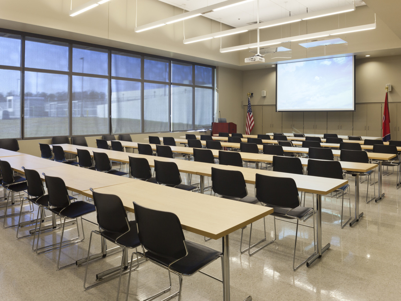 Long tables with chairs face podium and screen in beige room with large windows